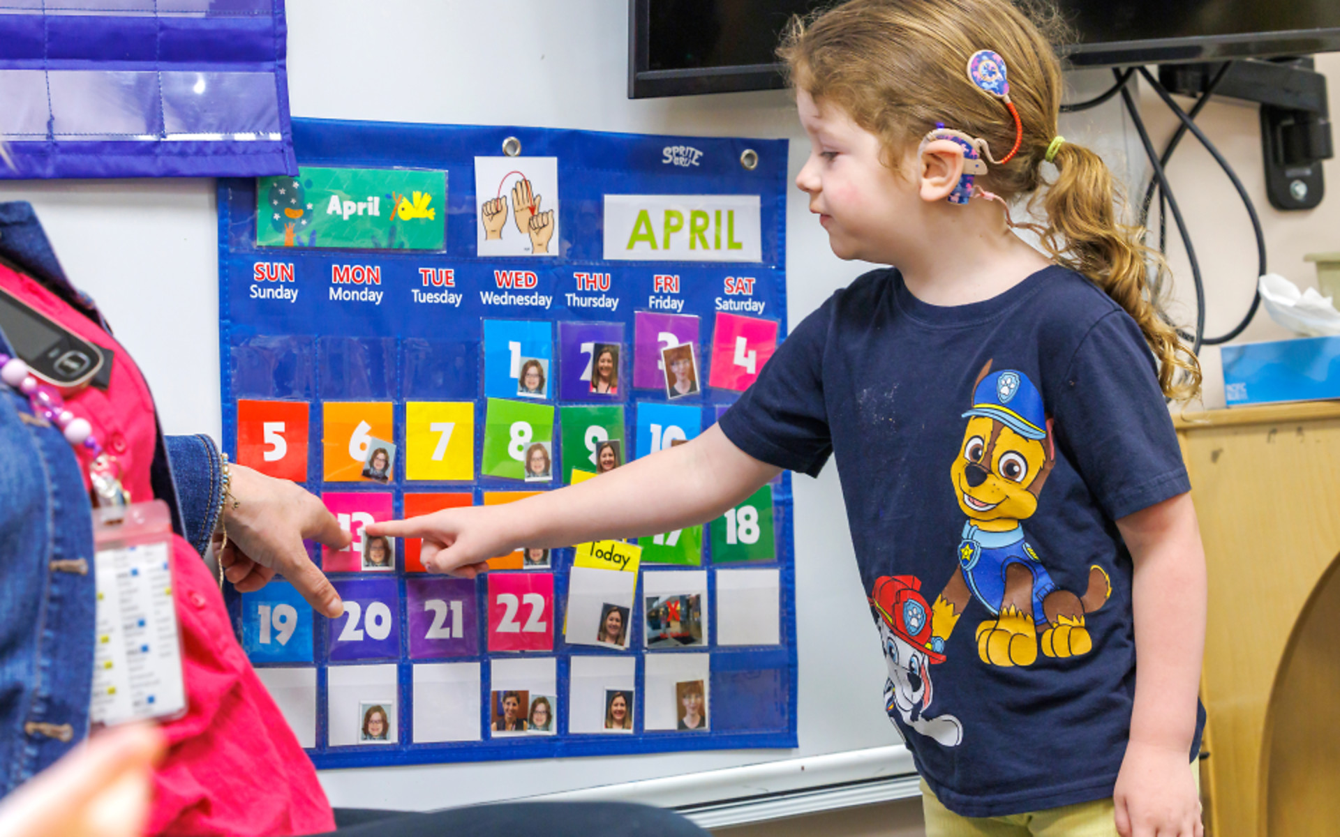 Young girl with hearing aids pointing to classroom calendar